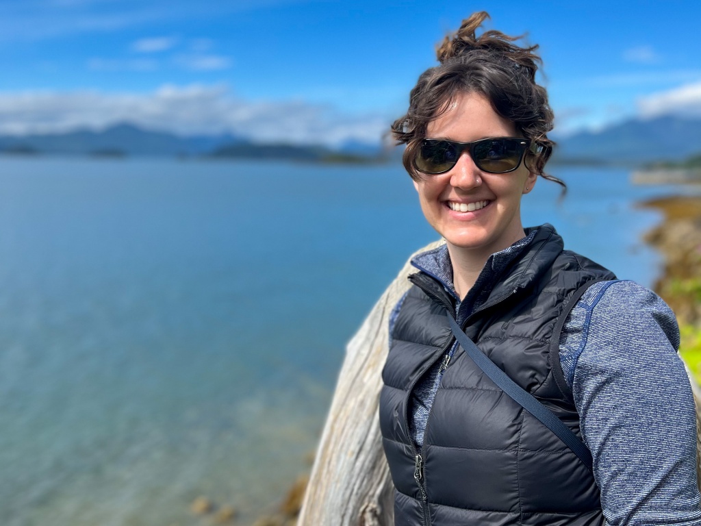 Kaitlyn smiling at the camera with a view of a body of water behind her and mountains in the distance.